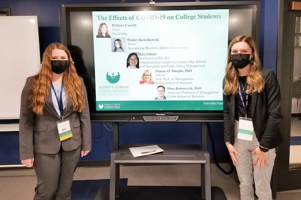 Two female students pose next to powerpoint presentation for SAM research group