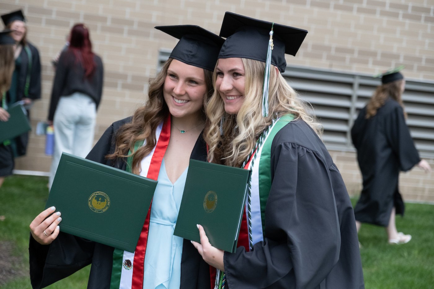 Two female students hold their diplomas and post for a photo outdoors after a Commencement ceremony.