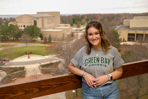 UW-Green Bay student smiles and poses with the campus in the background. Student is wearing a grey Green Bay Phoenix t-shirt.
