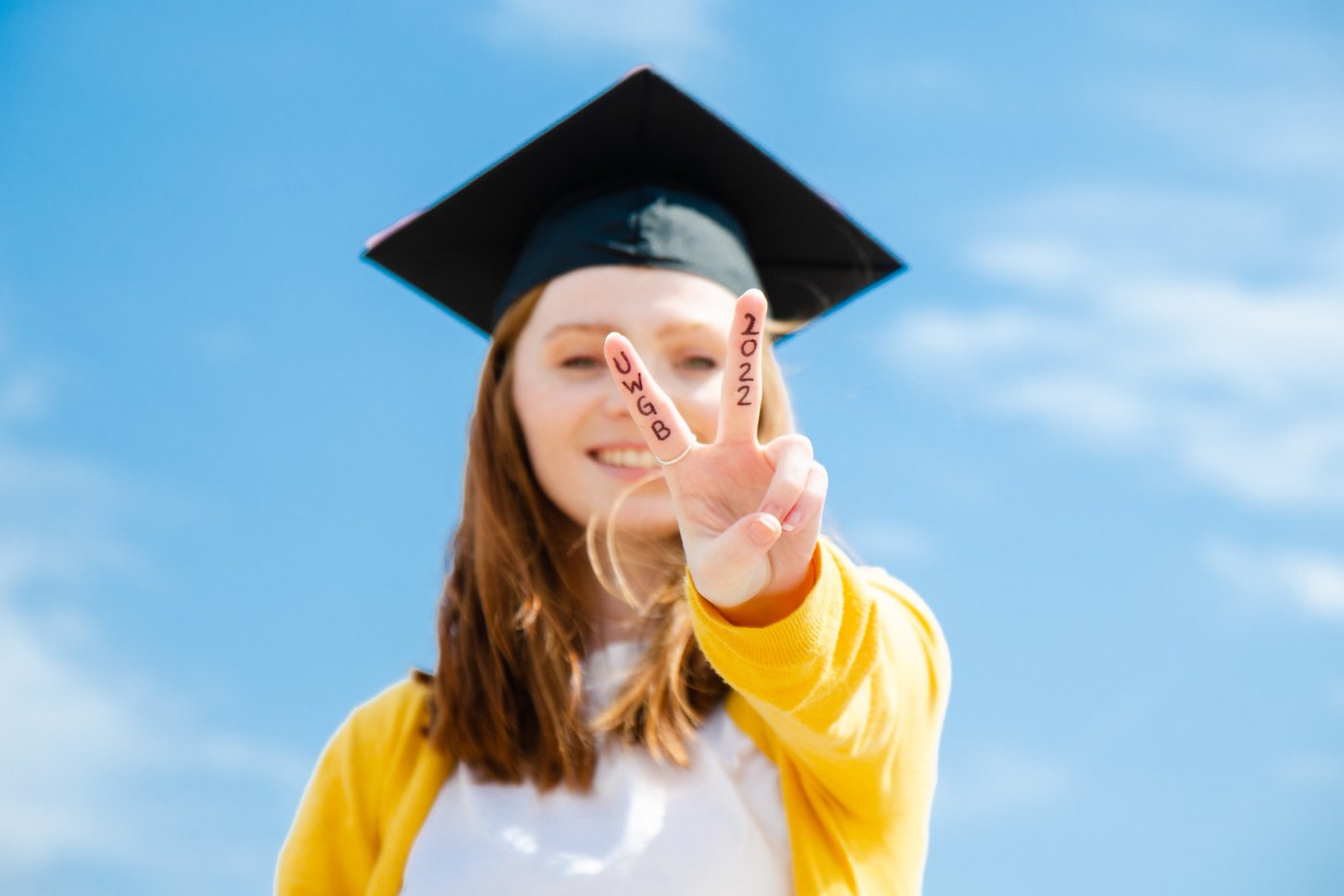 A female student posts in her grad cap with her fingers extended into a peace sign. The words "UWGB 2022" is written on her fingers.