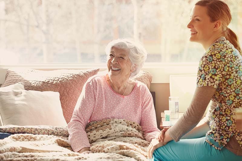 caregiver helping senior woman who is sitting in bed smiling