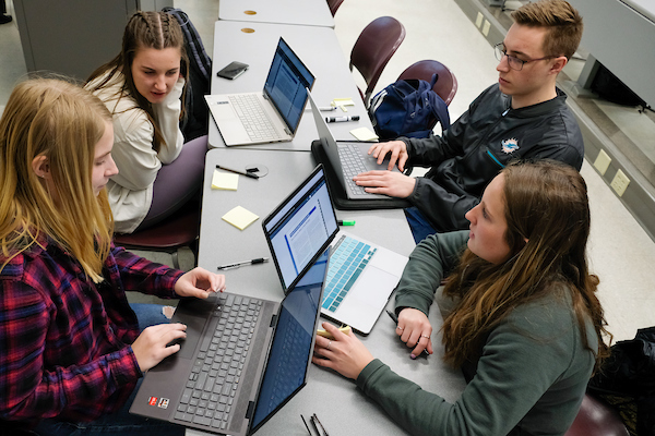 Four students work together in a small group during class. Two students are seated on each side of a long table. They are all working on their laptops and discussing the project.