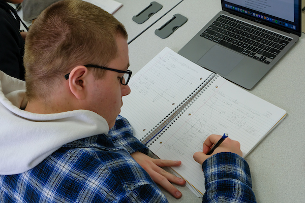 A student, wearing a blue plaid flannel and black frame glasses, is taking notes in calculus class. His laptop is open in front of him on the desk.