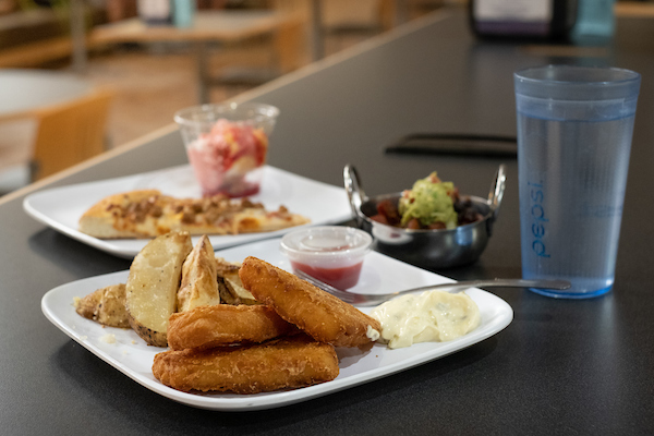 A variety of food, including pizza, potato wedges and fish, are arranged neatly on white plates on a table. There is also a small dish of another type of food topped with guacamole and a tall glass of water.