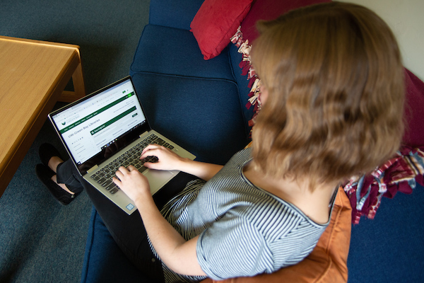 A student sits on a dark blue couch in their apartment-style residence hall room. They are browsing the UW-Green Bay website on their laptop.
