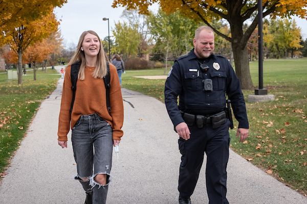 A student, wearing an orange sweater and dark jeans, walks alongside a university police offer outdoors on the UW-Green Bay campus.