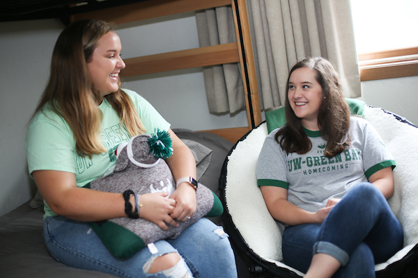 Two students sit together in a residence hall room. One student is sitting on the bottom bunk bed holding a UW-Green Bay pillow pet. The other student sits in a mushroom chair next to the bed. Both students are wearing UW-Green Bay apparel with blue jeans and are smiling at each other.