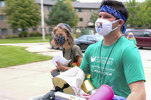 A move-in student volunteer walks alongside a new student moving into their residence hall. The volunteer is carrying a box of the student's belongings.