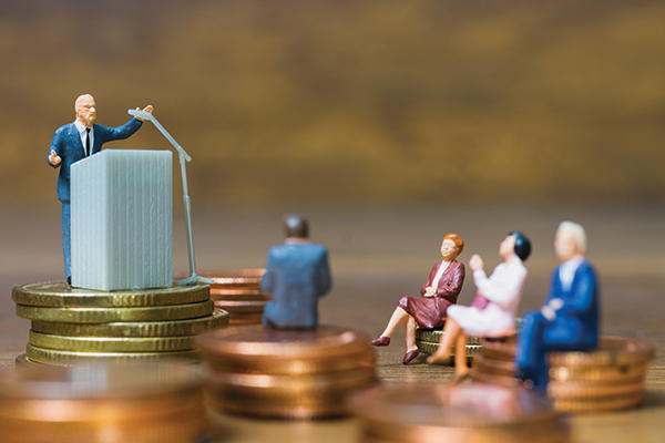 Small human figures sitting on coins facing a small podium.