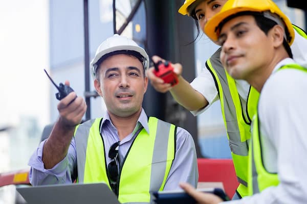 Three employees wearing hard hats look over shipping yard inventory