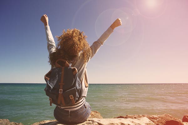 Young woman with a backpack, seated on a shoreline raising arms triumphantly