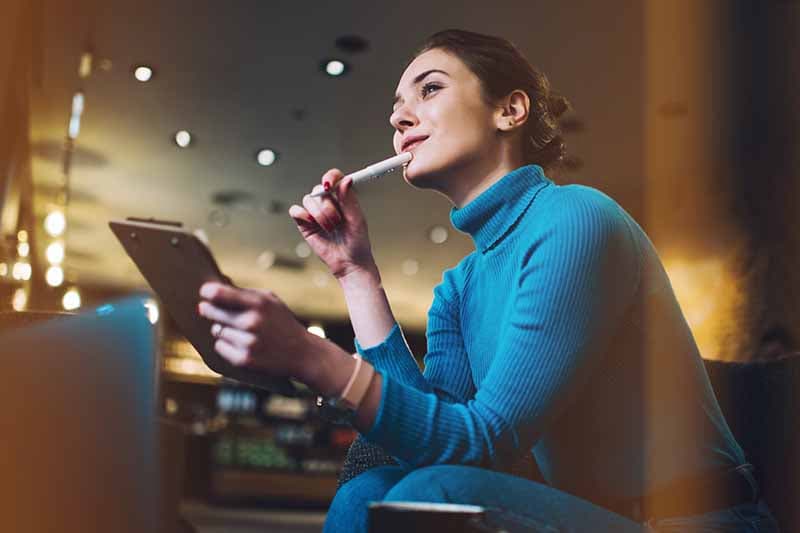 women in a blue turtleneck with a tablet holding a pen up to her mouth looking out into the distance