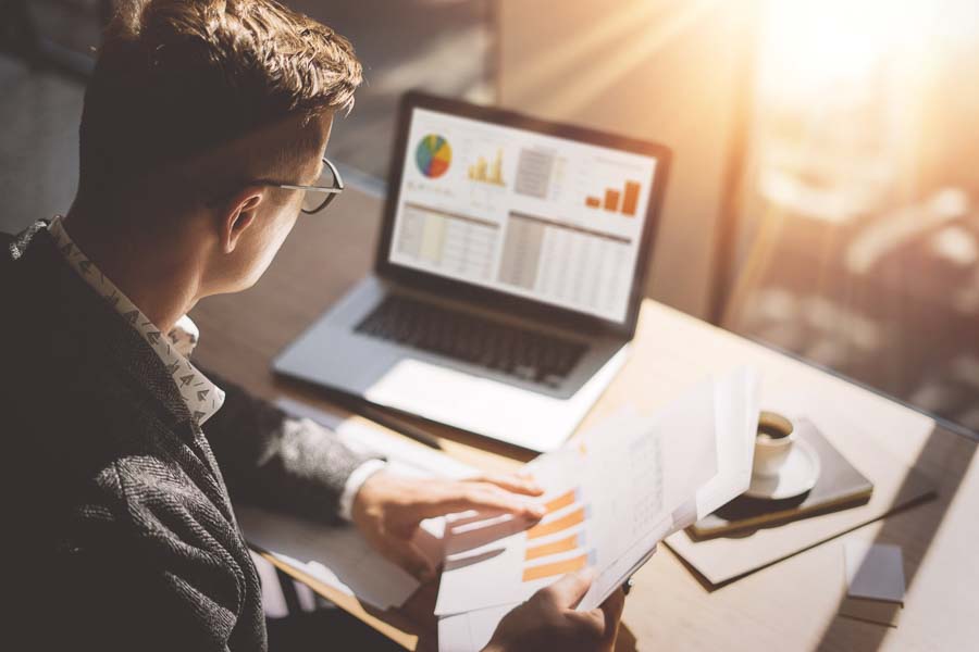 Young professional looking at printed charts and comparing it to a dashboard on a laptop screen