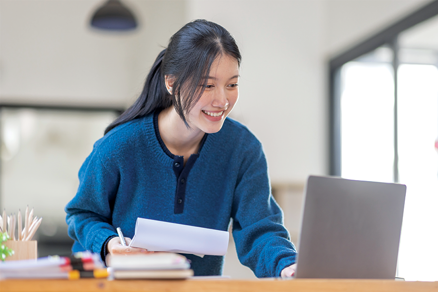 Woman holding pen and paper while typing on a laptop.