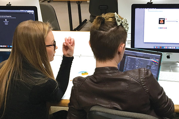 Two students look at each other's work on the Mac computers.