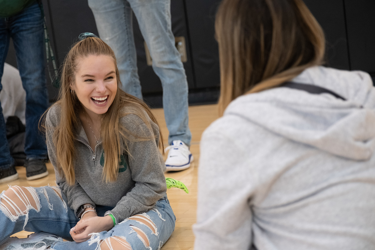 a student sits on the floor of the Kress Center gym during Krash the Kress. She is smiling at her friend who is sitting in the foreground.