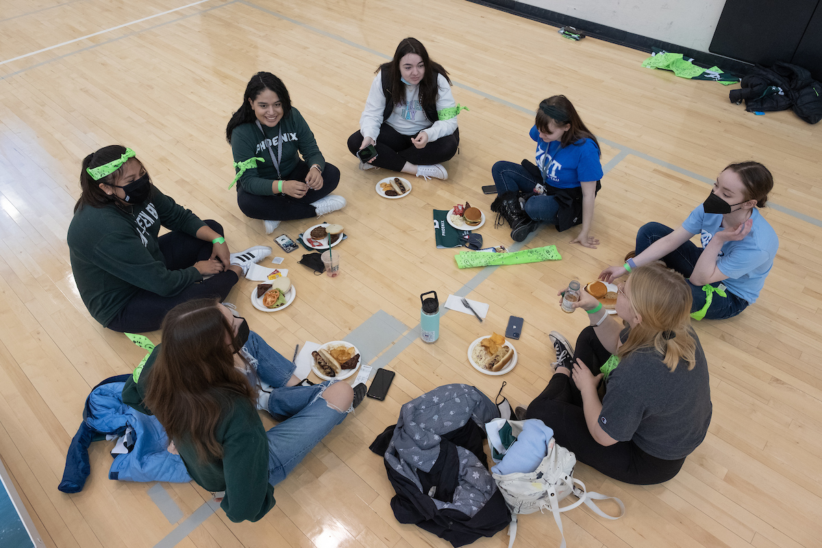 Seven students sit in a circle on the floor of the Kress Center gym and enjoy their free tailgate food.