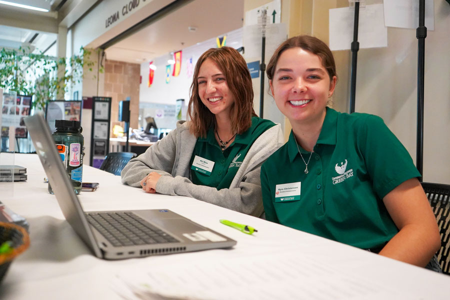 Two female university union employees working at desk smile for photo