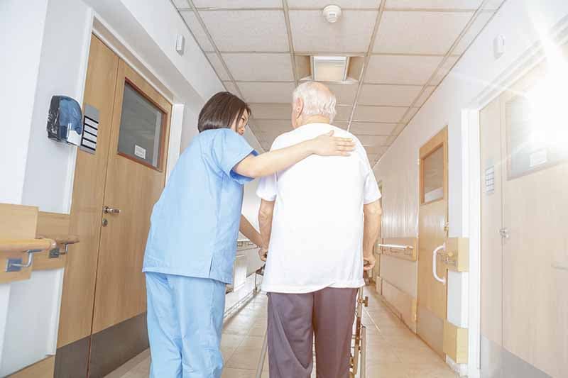 caregiver with hand on senior man's shoulder helping him walk in a hallway