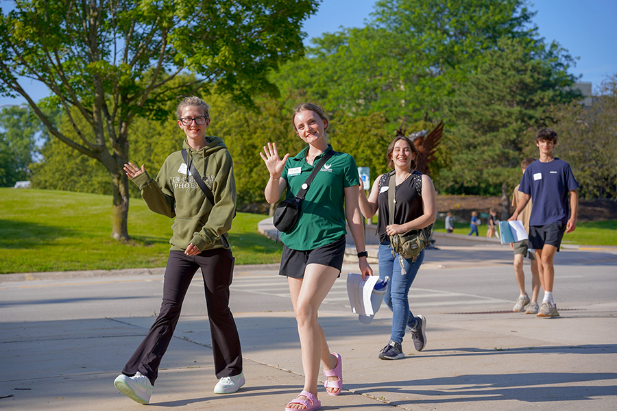 A student Ambassador and GB Orientation students walk on a campus sidewalk and wave at the camera