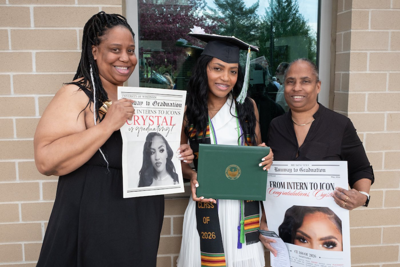 A graduated student stands with their diploma and two family members hold custom newspapers announcing their student's graduation