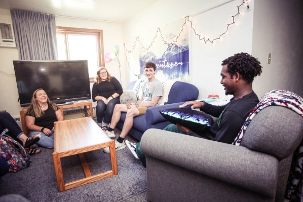Students sit around a table in a residence hall room. One student sits in a chair while speaking to the group, the others are focused on him.