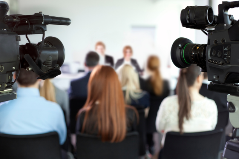 Media at a press conference being recorded by professional video cameras.