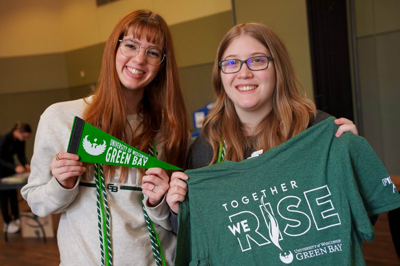 Students hold up a UW-Green Bay pennant (left) and a green "Together We Rise" t-shirt (right)