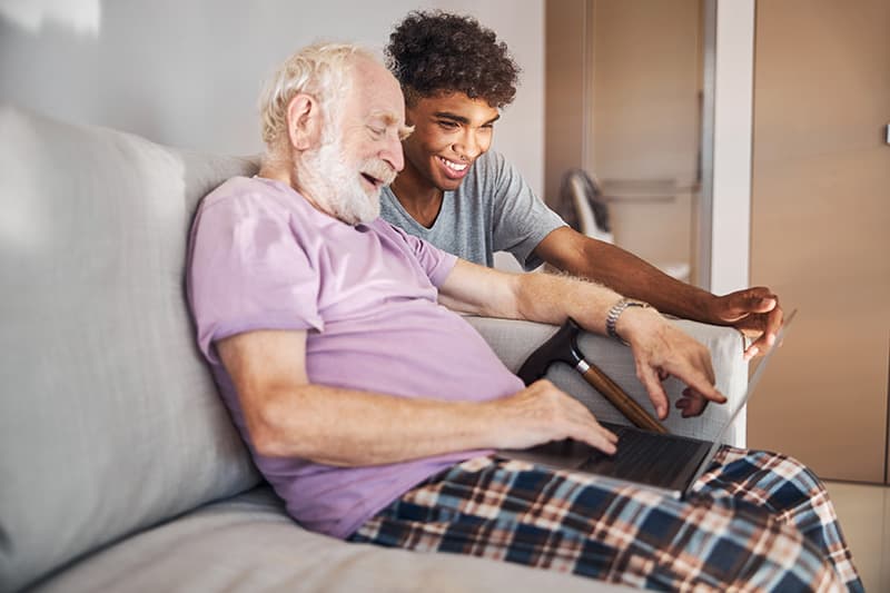 young man caregiver sitting on couch helping senior man with laptop