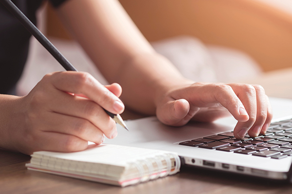 A person at a laptop taking notes on a small notepad