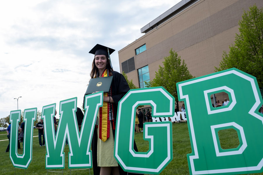 Student in graduation cap and gown posing by UW-Green Bay sign