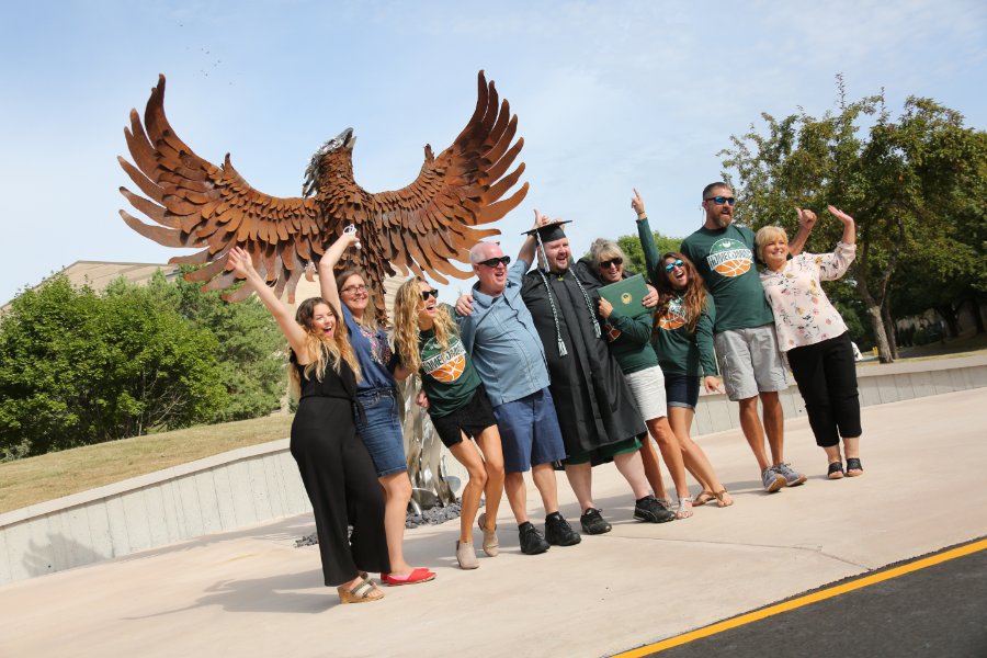 Family and friends surround a recent UW-Green Bay graduate. They are posed in front of the Phoenix sculpture smiling and celebrating for the camera.