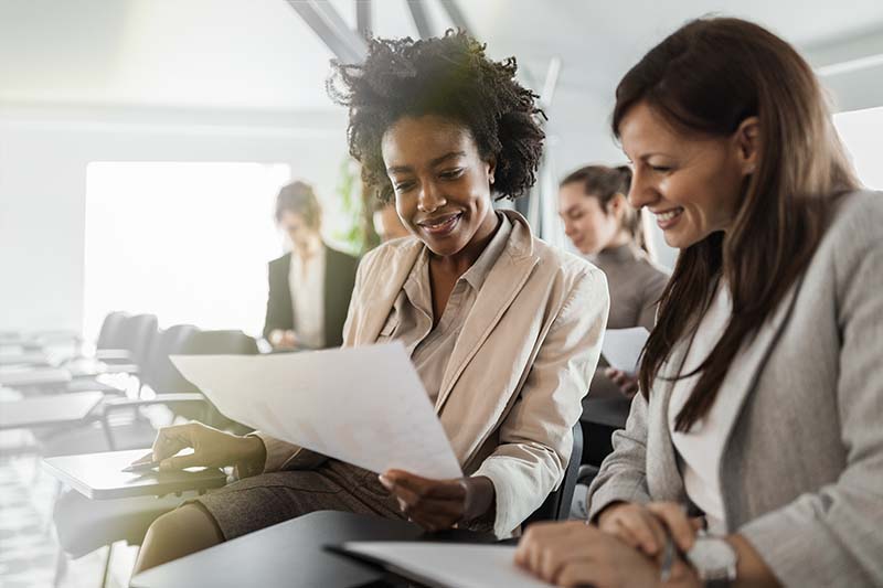 two women dressed in suits smiling looking at a worksheet during class