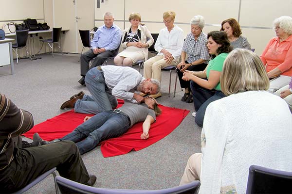 Participants seated in a semi-circle observe a First Aid CPR demonstration.