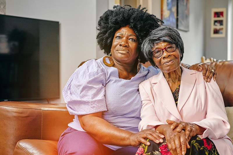 woman caregiver sitting on couch with her hand on senior woman's shoulder