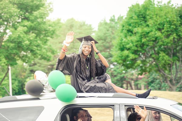 Black female graduate sits on top of decorated car for commencement ceremony procession.