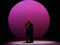 Two actors embracing on stage with a large pink spotlight on them