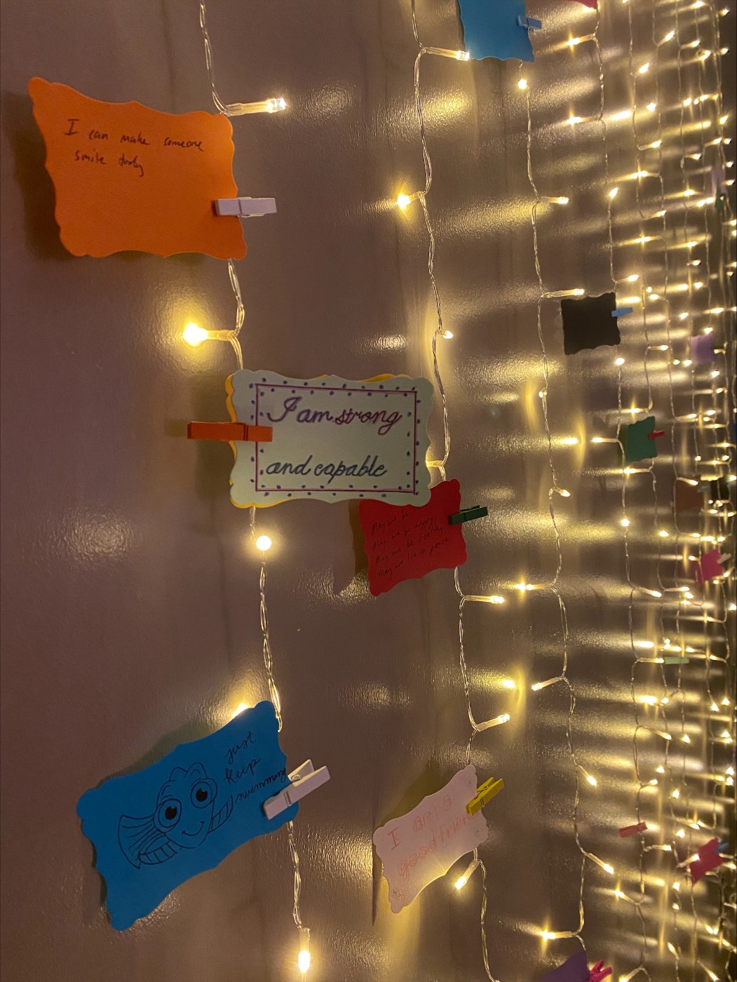 Close up image of note cards with positive messaging close pinned to string lights along a wall in a dark room.