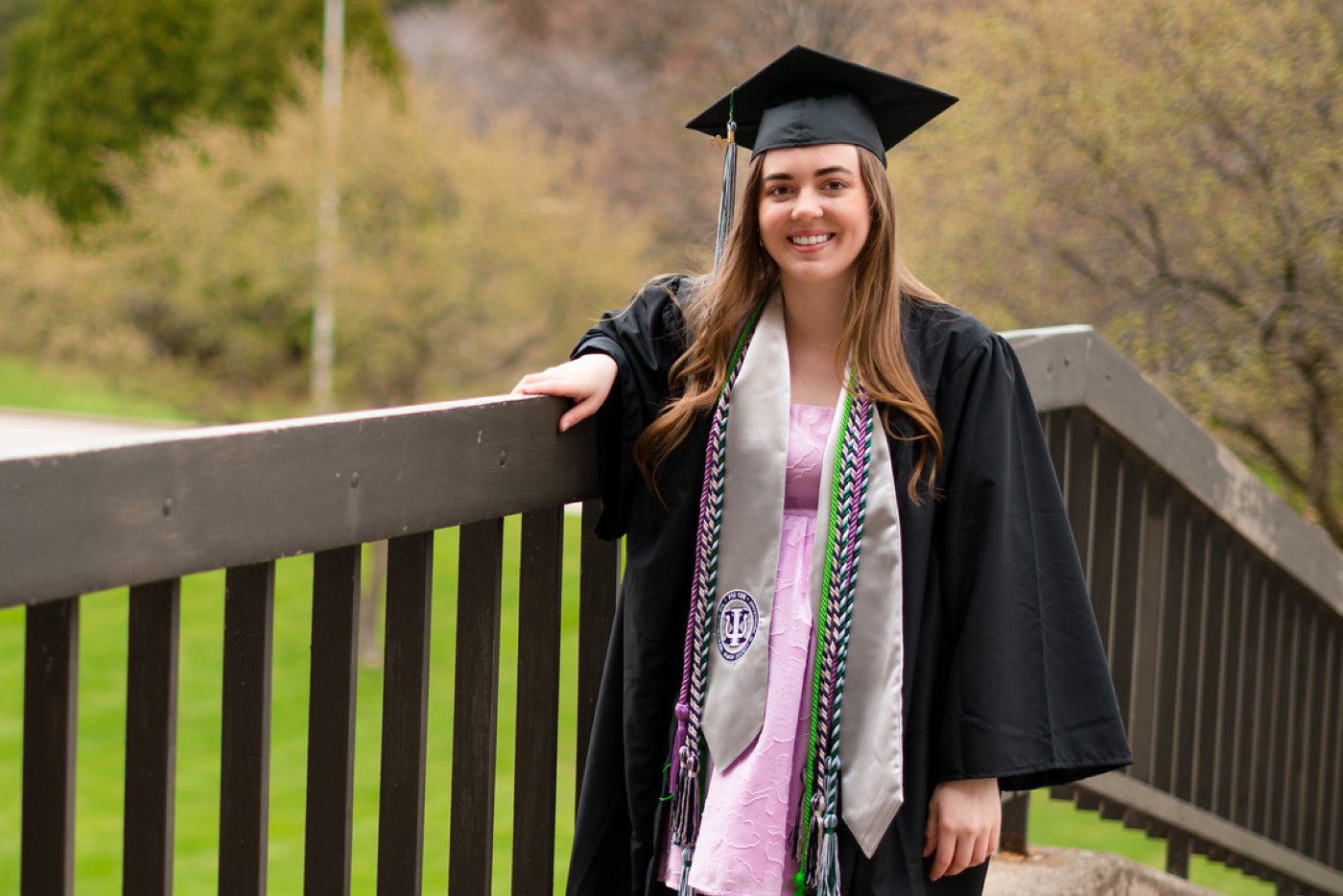 A graduating student poses for a photo in their cap, gown, cords and other regalia