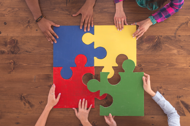 A background of a table with five hands each holding a piece of a large puzzle in different colors (yellow, blue, green, red) coming together