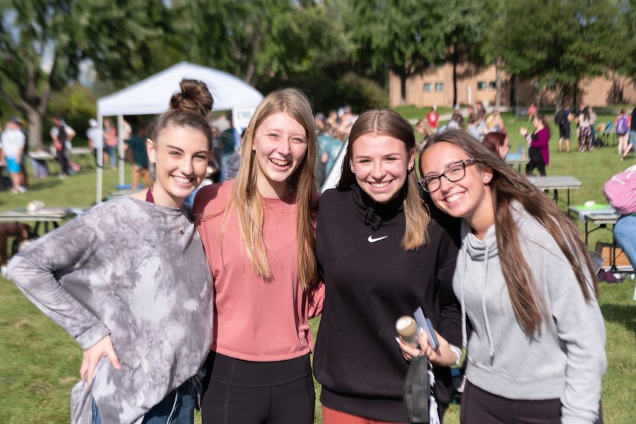 Four students stand in a line with their arms around each other. They are smiling for the camera, and OrgSmorg activities can be seen in the background behind them.