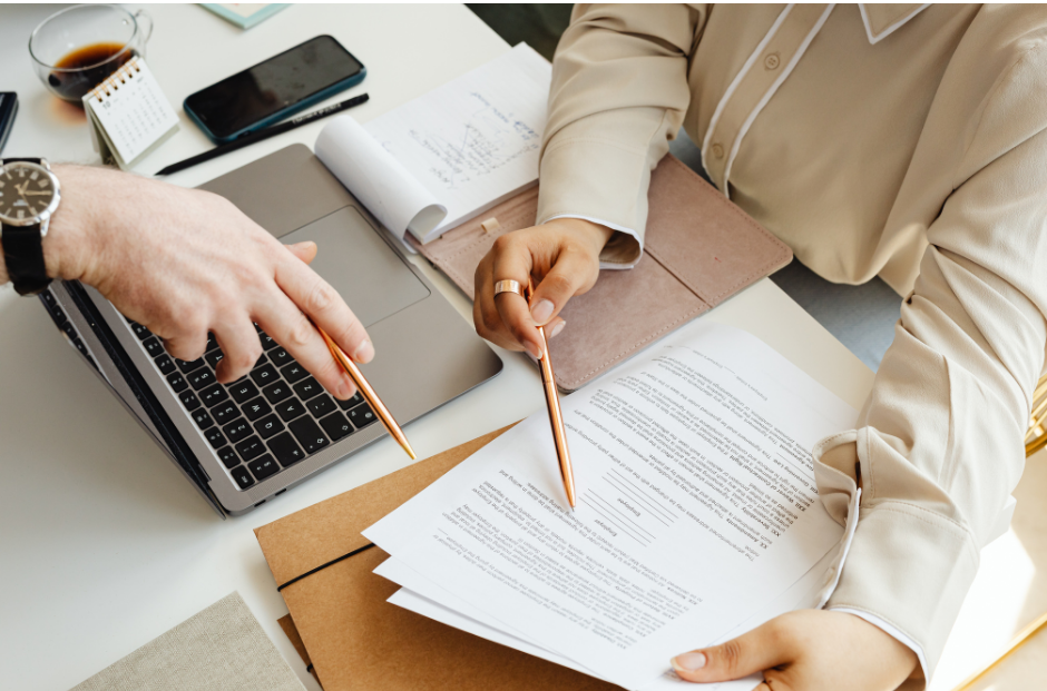 Two people reviewing printed data with a laptop to the left of the paperwork