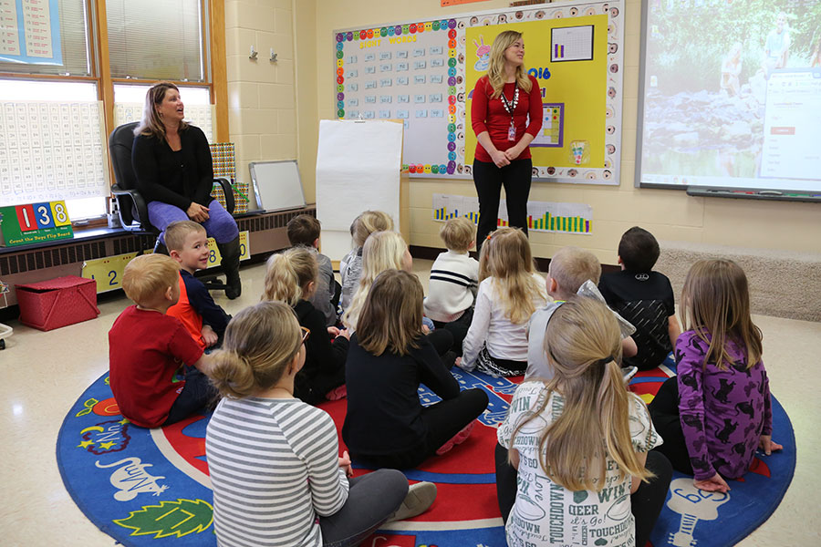 Teacher standing in front of a group of children in a classroom looking at a projected image on a whiteboard..