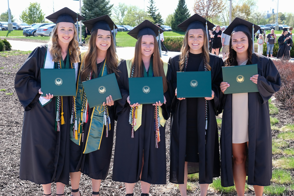 Five spring 2022 graduates pose in their caps, gowns, honors cords and regalia while holding their diplomas.