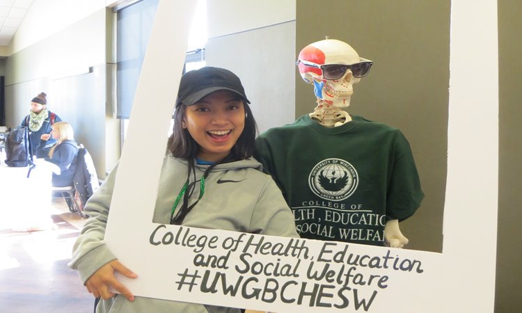 Student holding a #UWGBCHESW frame around her and a skeleton with a UWGB shirt on
