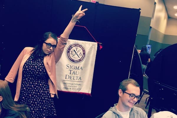 Writing and Applied arts student poses with the peace sign in front of Sigma Tau Delta logo