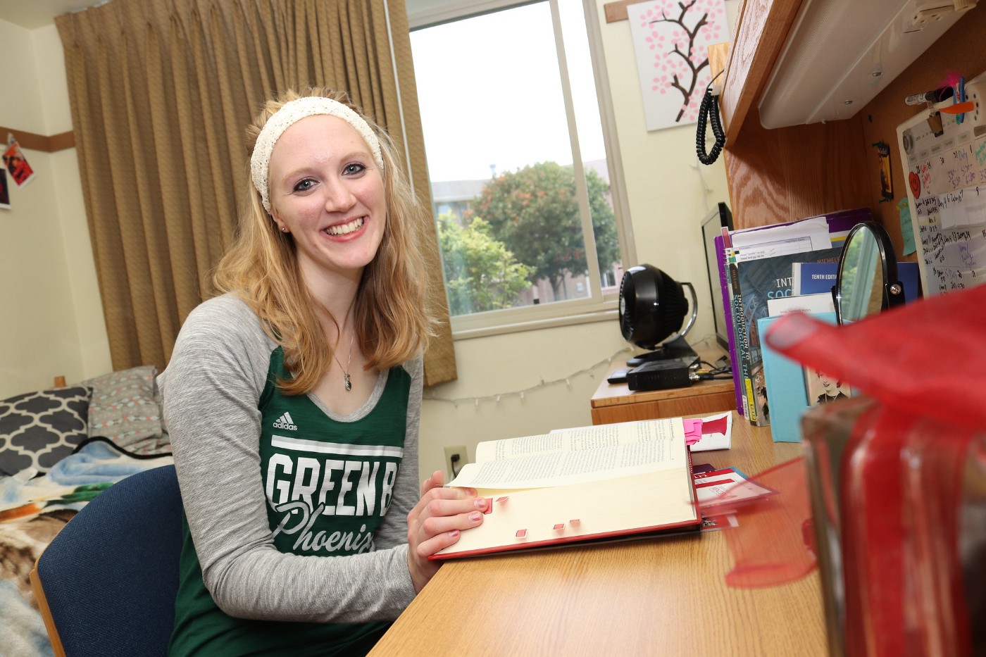 A student sits at a desk in her dorm room with a large textbook in front of her.