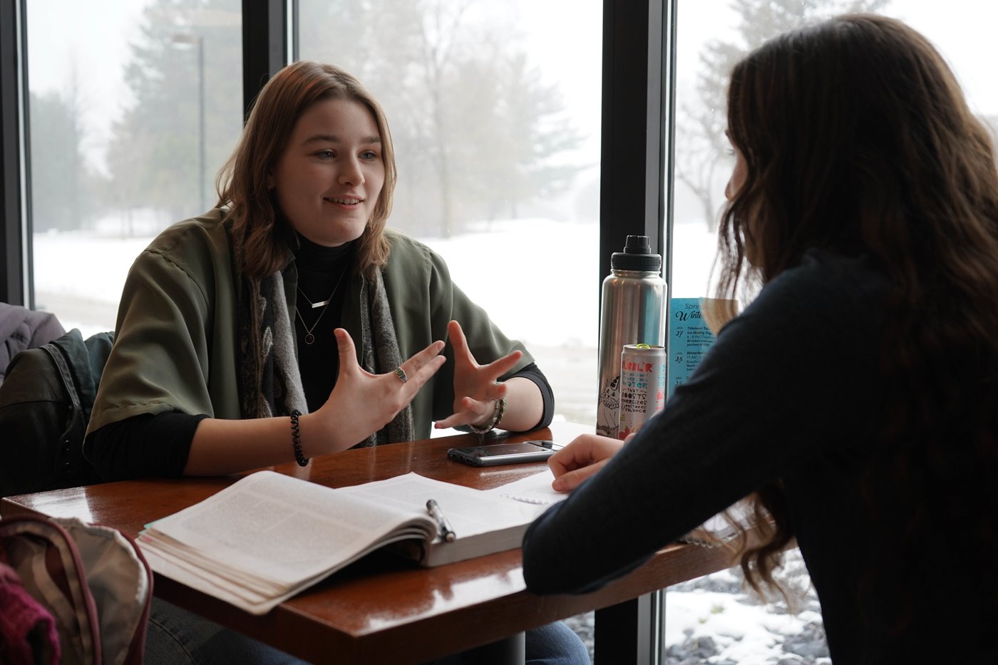 Two students sit at a table in front of a window with textbooks and water bottles between them.