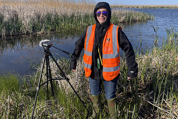 Haley Spargur, wearing rubber boots standing in marsh grass beside a tripod with recording equipment